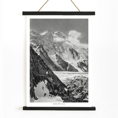 Black and white photograph and poster of Broad Peak by Vittorio Sella showing snow covered mountains and mountaineers climbing amidst rocky terrain and glacier, with wooden magnetic frame on white background