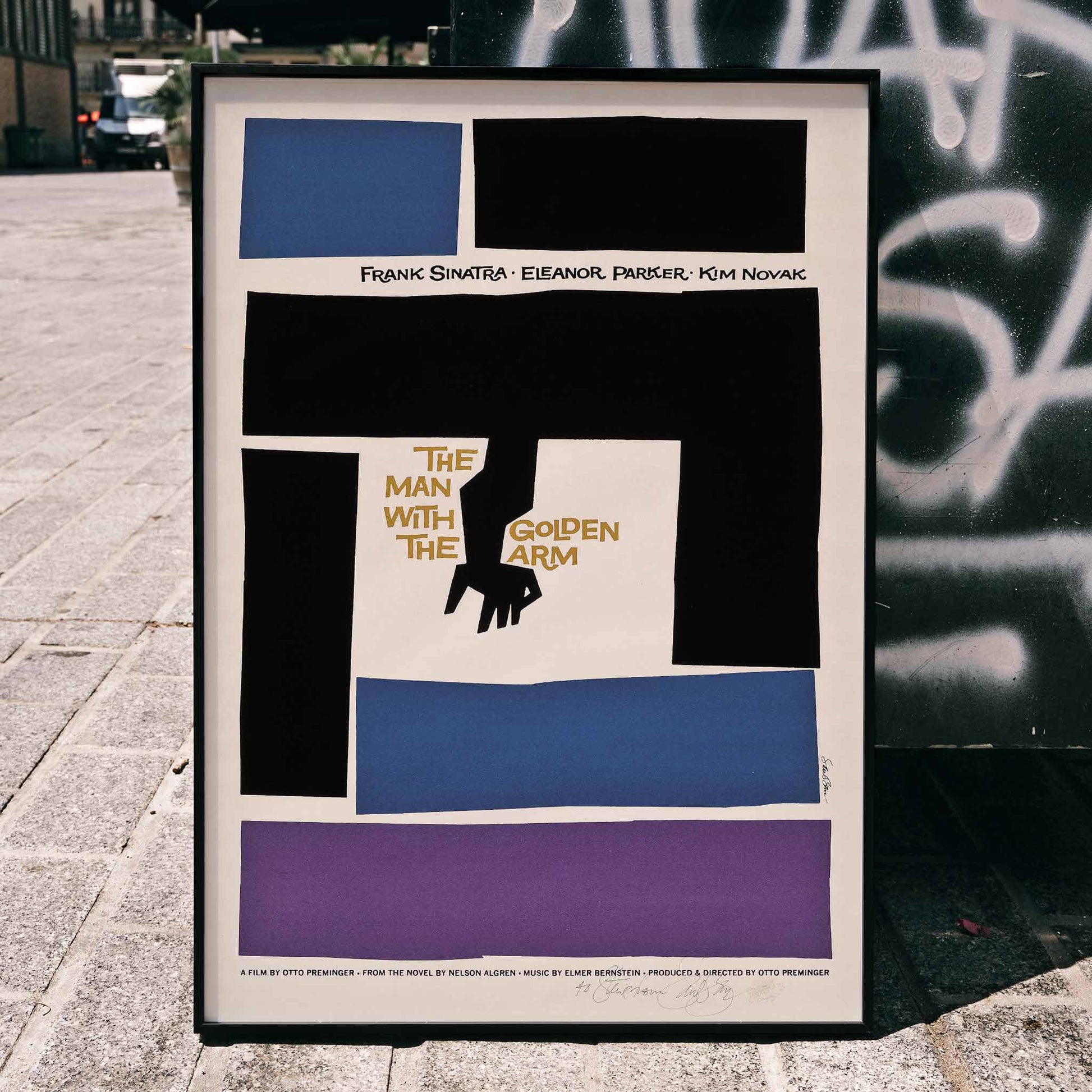 Movie poster for The Man with the Golden Arm by Saul Bass featuring bold abstract shapes and a stylized arm and hand in vibrant colors, framed and displayed in home decor