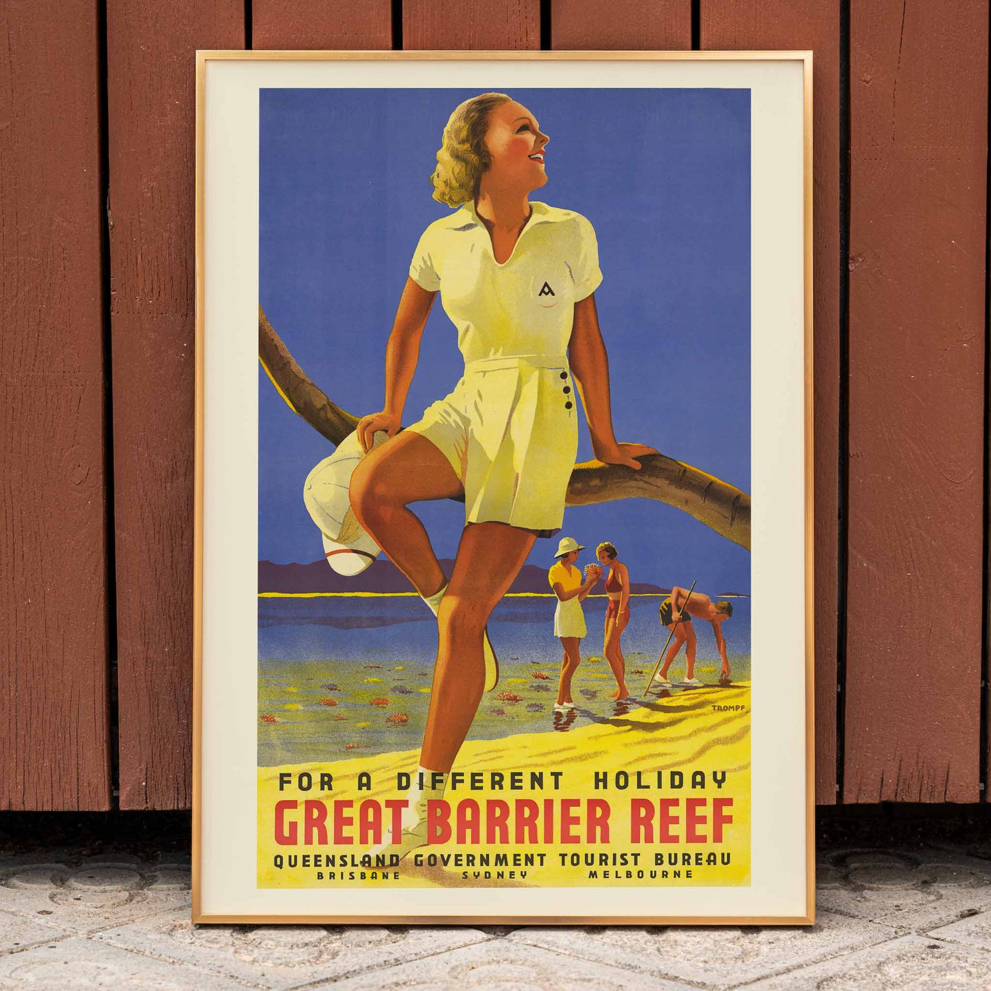 Vintage travel poster by Percival Albert Trompf showing a woman sitting on a branch at the Great Barrier Reef with people enjoying the beach in the background, framed and displayed in home decor